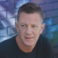 Man with short dark hair and blue eyes against brick wall.